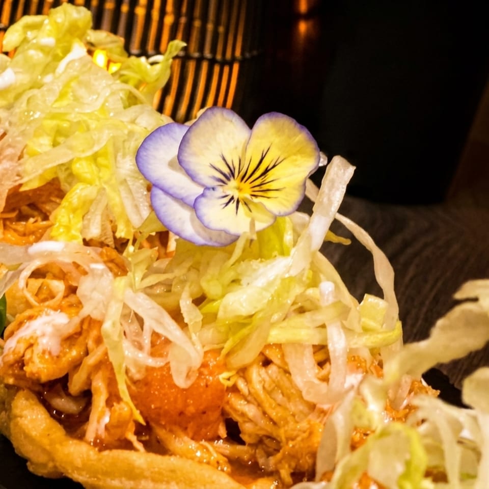 Two sopes topped with shredded chicken, lettuce, crema, and a purple and yellow edible flower.