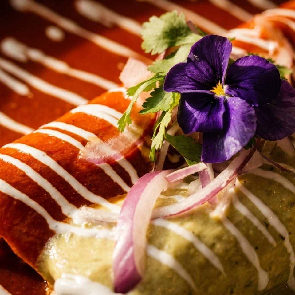 Two enchiladas, one red and one green, drizzled with crema and garnished with a purple flower.