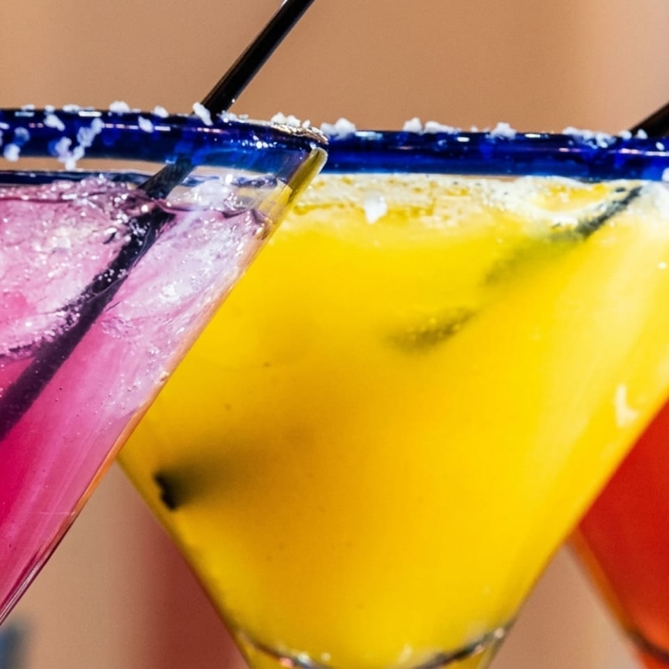 Three vibrant margaritas in blue-rimmed glasses: pink, yellow, and red, with straws and salt.