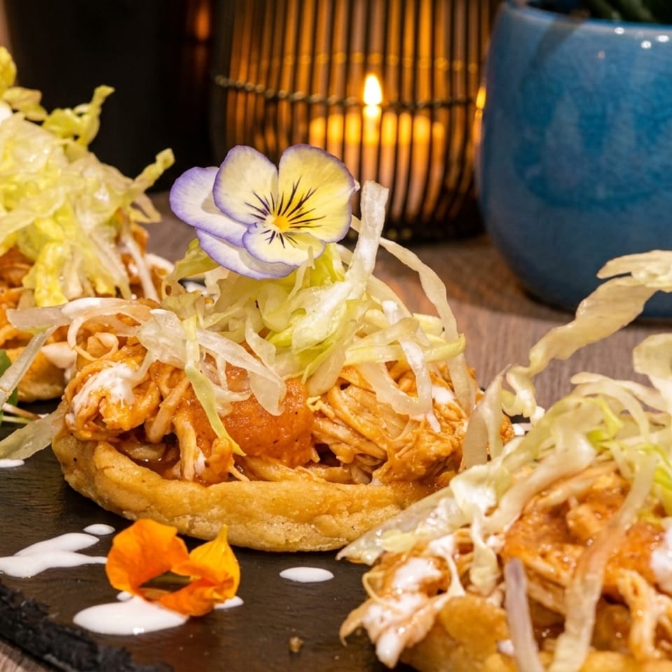 Three chicken sopes topped with shredded lettuce, cream, and edible flowers on a dark slate.